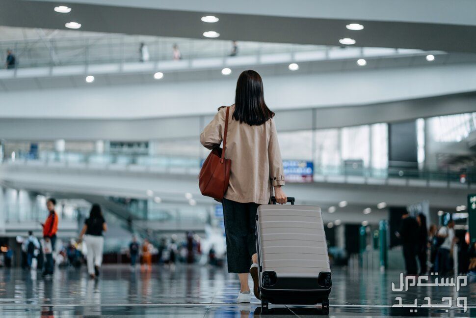 قطع تذاكر طيران باسعار رخيصة واحيانا مجانية! كيف؟ فتاة تقف بظهرها في المطار وتجر حقيبة سفر وراءها