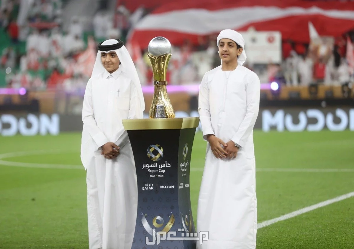 مواعيد مباريات السوبر الإماراتي القطري والفرق المشاركة صورة لطفلين يقفان إلى جانب كأس السوبر الإماراتي القطري